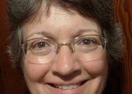 Professional headshot of Christian life coach minister Elizabeth smiling while wearing glasses against a wood-paneled background.