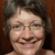 Professional headshot of Christian life coach minister Elizabeth smiling while wearing glasses against a wood-paneled background.