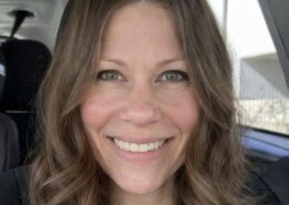 A smiling woman with wavy brown hair sits in a car, looking at the camera.