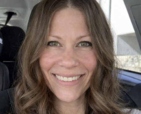 A smiling woman with wavy brown hair sits in a car, looking at the camera.