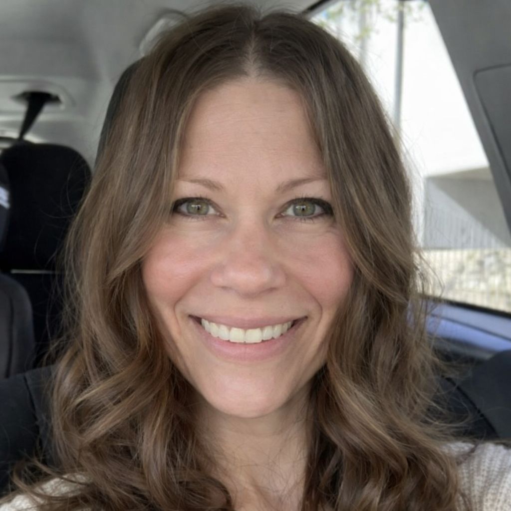 A smiling woman with wavy brown hair sits in a car, looking at the camera.