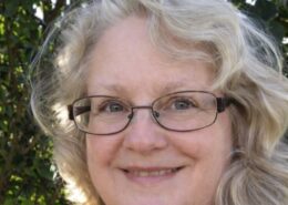 A smiling woman with glasses and light hair stands outdoors in front of leafy greenery.