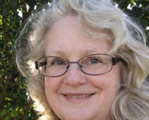 A smiling woman with glasses and light hair stands outdoors in front of leafy greenery.