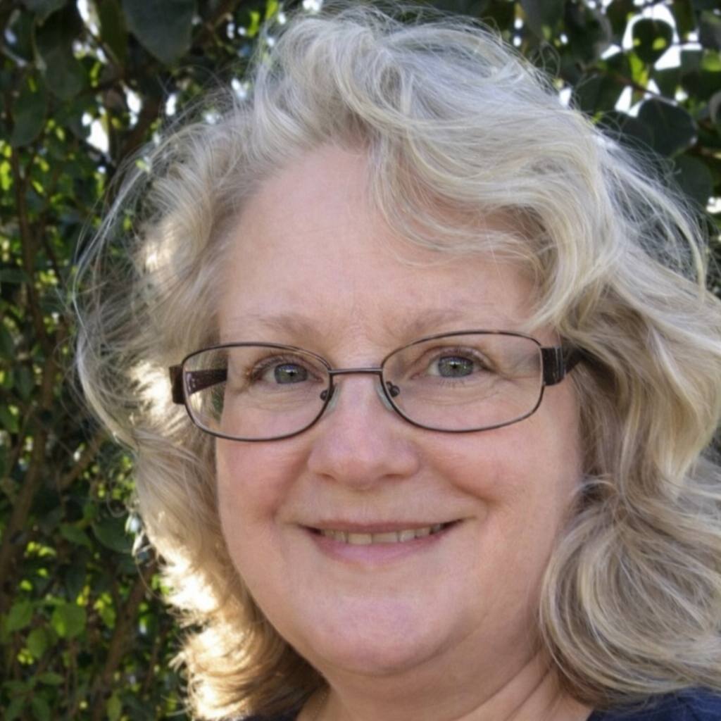 A smiling woman with glasses and light hair stands outdoors in front of leafy greenery.