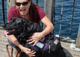 A smiling woman wearing sunglasses hugs a black dog in a purple service vest while sitting on a wooden dock by the water.