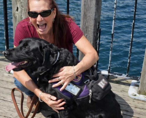 A smiling woman wearing sunglasses hugs a black dog in a purple service vest while sitting on a wooden dock by the water.