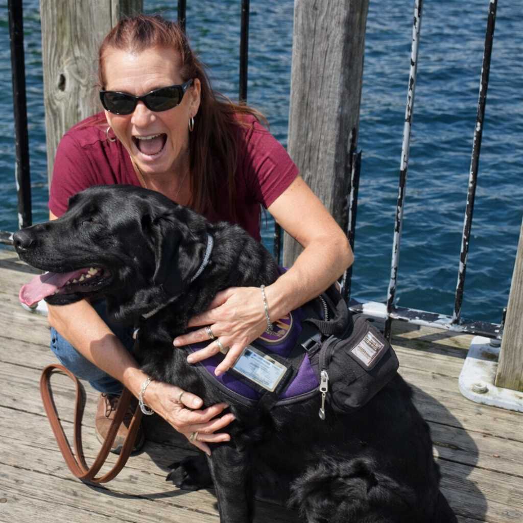 A smiling woman wearing sunglasses hugs a black dog in a purple service vest while sitting on a wooden dock by the water.