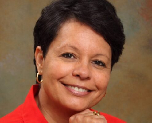 A smiling woman with short dark hair rests her chin on her hand in a studio-style portrait.