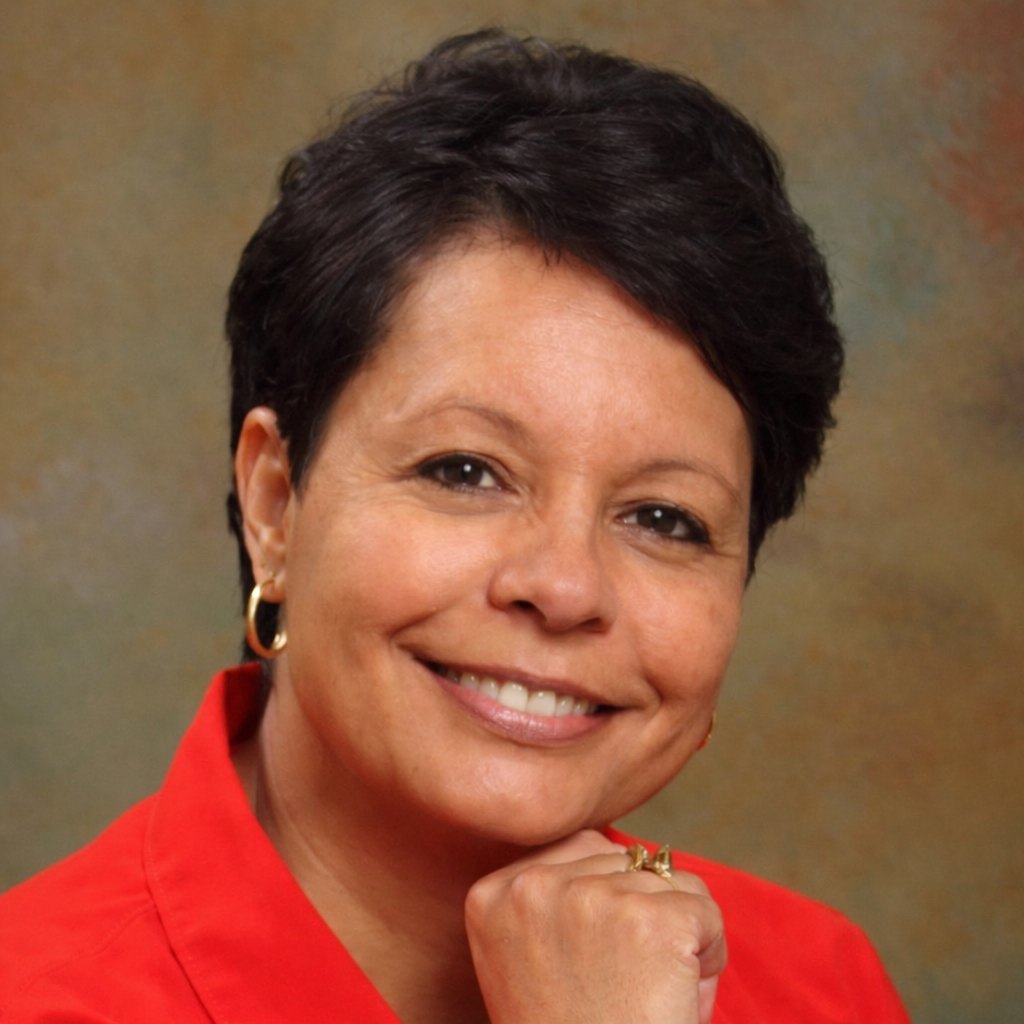 A smiling woman with short dark hair rests her chin on her hand in a studio-style portrait.