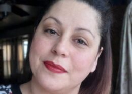 A woman with dark hair and red lipstick looks at the camera in a softly lit indoor setting.