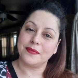 A woman with dark hair and red lipstick looks at the camera in a softly lit indoor setting.