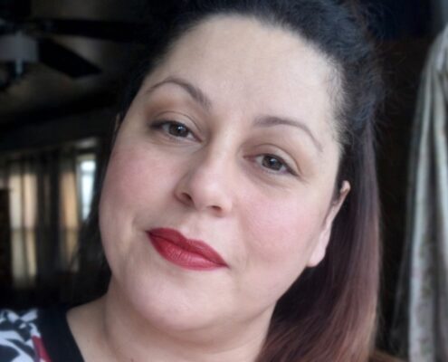 A woman with dark hair and red lipstick looks at the camera in a softly lit indoor setting.
