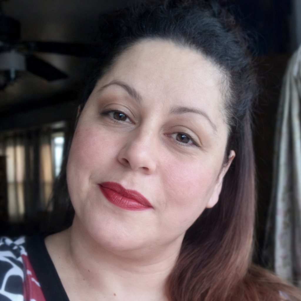 A woman with dark hair and red lipstick looks at the camera in a softly lit indoor setting.