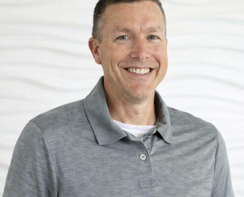 A smiling man in a gray polo shirt poses against a light, neutral background.