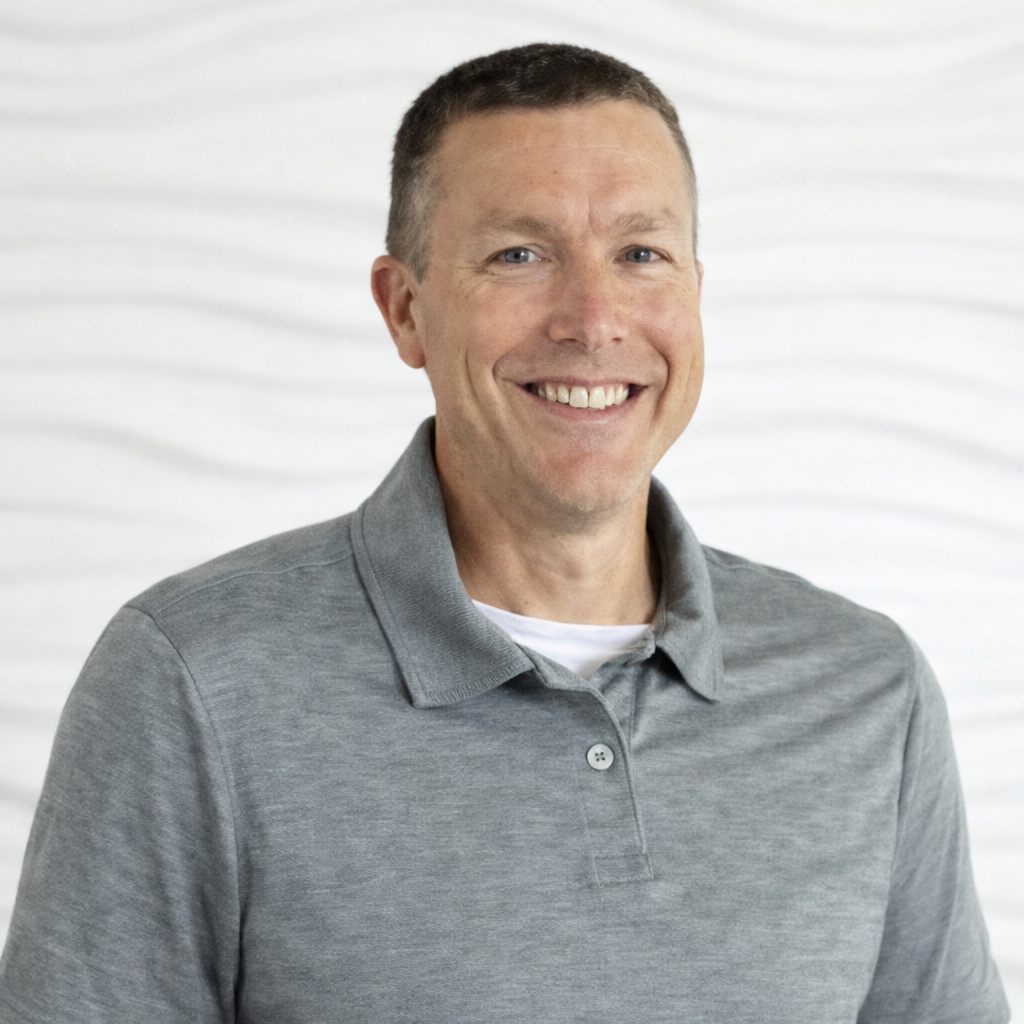 A smiling man in a gray polo shirt poses against a light, neutral background.