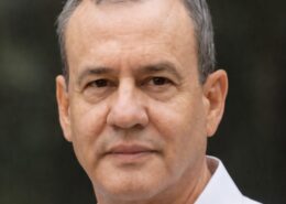Close-up portrait of a middle-aged man with short gray hair wearing a light-colored collared shirt, looking at the camera against a blurred green background.