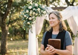 Female Christian wedding officiant holding a Bible beside a floral wedding arch during an outdoor wedding ceremony.