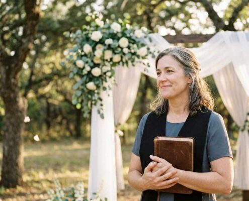 Female Christian wedding officiant holding a Bible beside a floral wedding arch during an outdoor wedding ceremony.