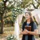 Female Christian wedding officiant holding a Bible beside a floral wedding arch during an outdoor wedding ceremony.