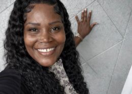 A smiling woman with long curly hair takes a selfie indoors, standing beside a textured wall.