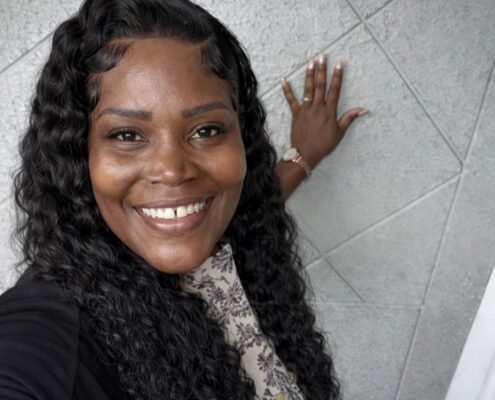 A smiling woman with long curly hair takes a selfie indoors, standing beside a textured wall.