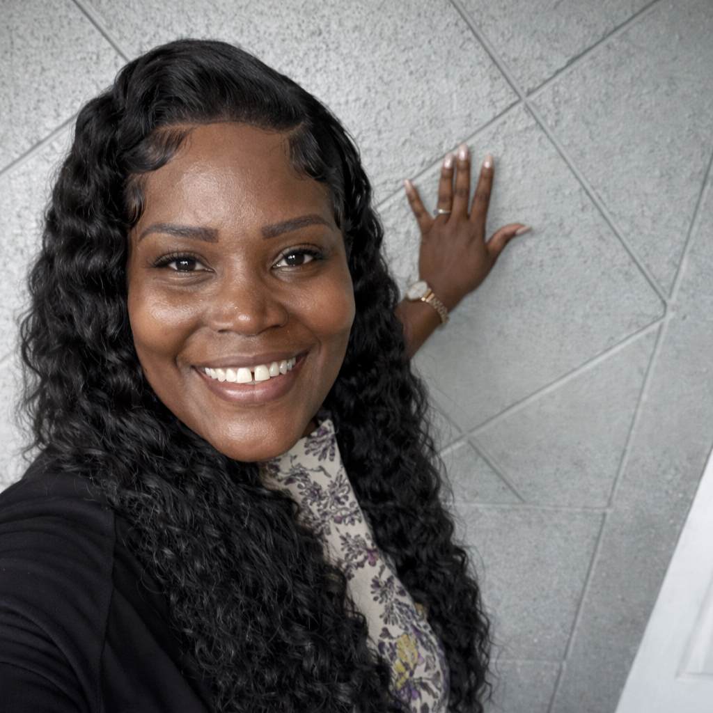 A smiling woman with long curly hair takes a selfie indoors, standing beside a textured wall.