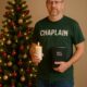 Casual Christmas Chaplain in t-shirt and jeans holding a Bible and candle beside a decorated Christmas tree, symbolizing prayer and ministry during the holiday season.