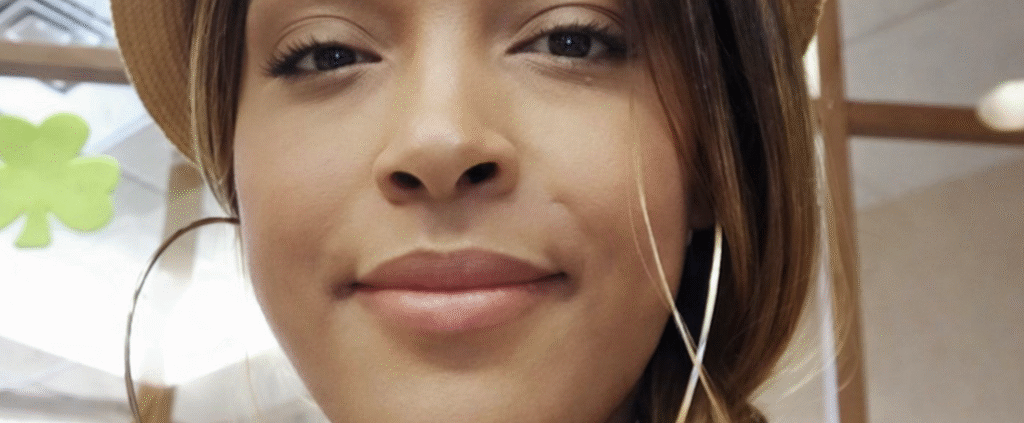 Close-up portrait of a woman wearing a tan hat and large hoop earrings, with her hair styled in a side braid, looking calmly at the camera in an indoor setting.