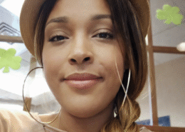 Close-up portrait of a woman wearing a tan hat and large hoop earrings, with her hair styled in a side braid, looking calmly at the camera in an indoor setting.