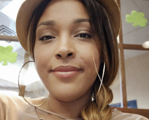 Close-up portrait of a woman wearing a tan hat and large hoop earrings, with her hair styled in a side braid, looking calmly at the camera in an indoor setting.