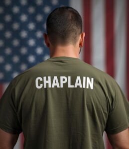 A person wearing a shirt labeled “CHAPLAIN” stands facing an American flag.