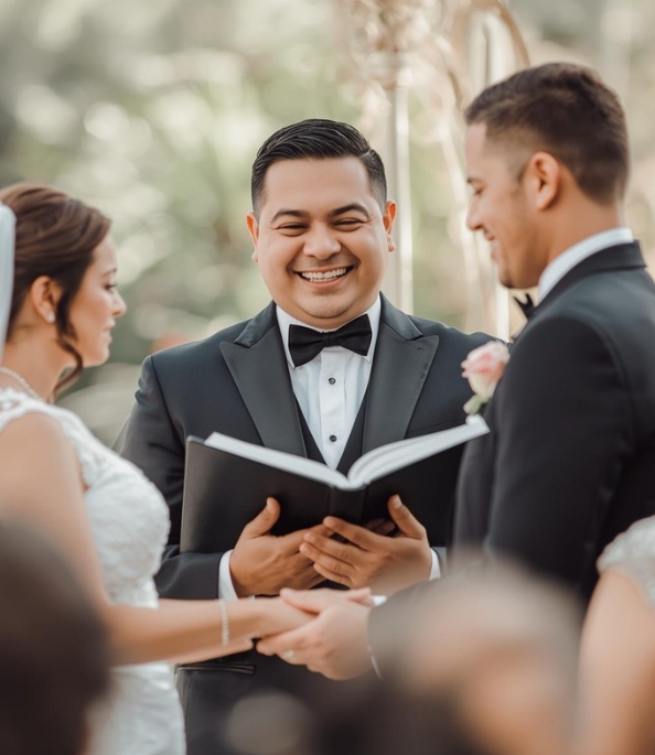 A smiling wedding officiant holds an open book while standing between a bride and groom during a ceremony.
