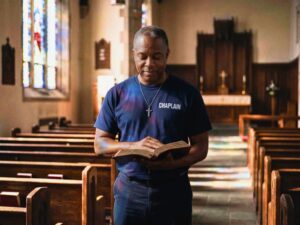 Male chaplain wearing CHAPLAIN shirt praying while holding Bible.