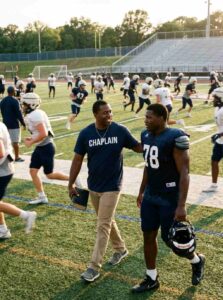 Black male sports chaplain encouraging football players during practice while holding a Bible and wearing a CHAPLAIN shirt.