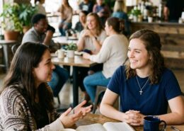 Young Christian woman wearing a navy blue t-shirt and cross necklace listening attentively during a Bible-centered conversation with another woman in a welcoming church café.